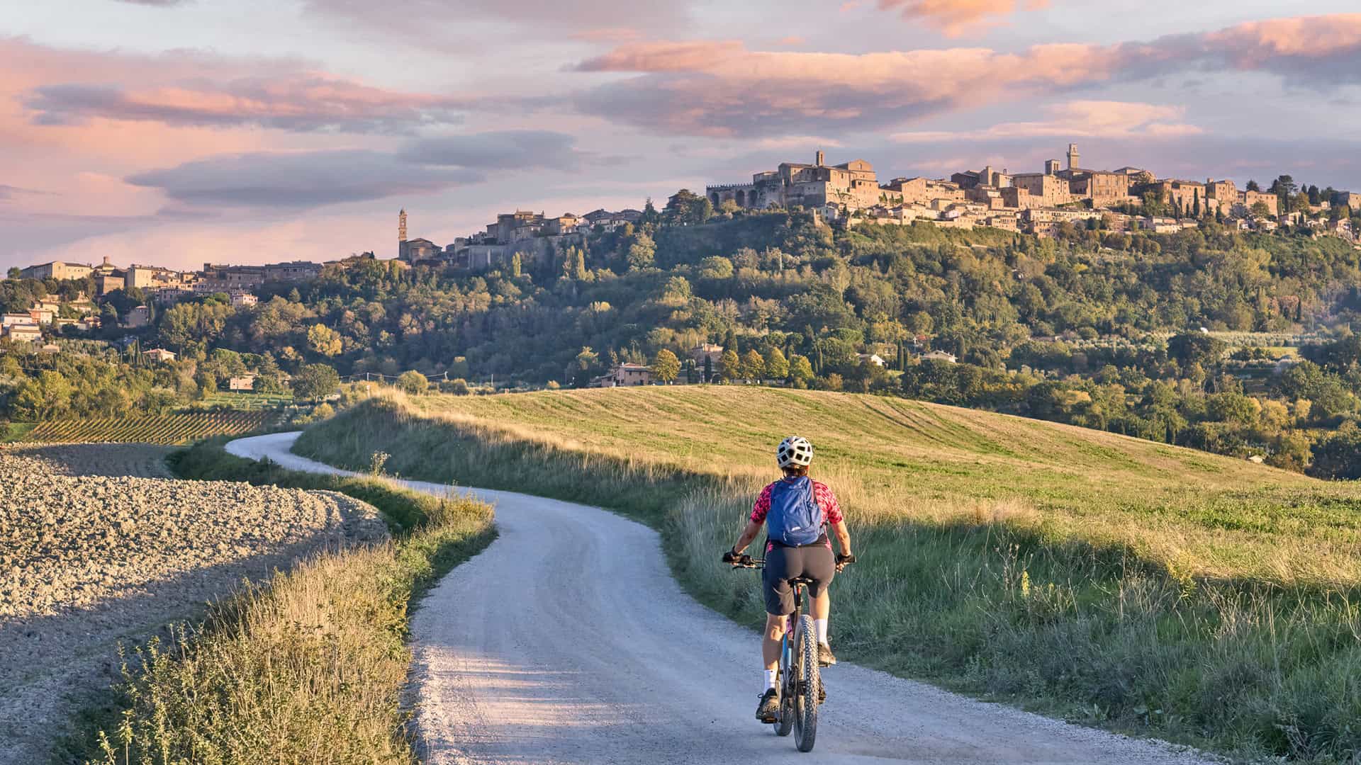 bella donna anziana in sella alla sua mountain bike elettrica tra gli ulivi nella zona del Chianti con la città medievale di Montepulciano sullo sfondo, Toscana, Italia